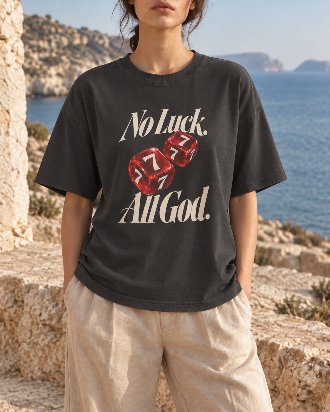 No Luck, All God Boxy T-Shirt — Vintage Black. AI-created editorial styling image beside sunlit stone and the sea.
